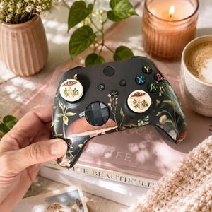 May include: A black wireless video game controller featuring a botanical mushroom and foliage print. The controller includes two white thumbstick covers with a small mushroom illustration, a black directional pad, and standard colored action buttons.