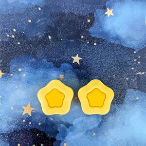 May include: Two yellow star-shaped earrings with a smaller yellow star inside each. The earrings are on a blue background with white stars.