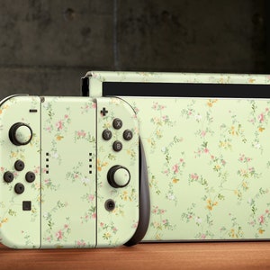 Pode incluir: Um console e um controlador Nintendo Switch verdes com um padrão floral. O console e o controlador estão em uma superfície de madeira.