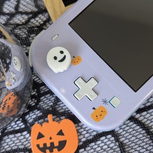 Pu&ograve; includere: Una console portatile color viola chiaro con un copri-joystick bianco a forma di fantasma, piccoli adesivi a forma di zucca arancione e un pad direzionale. Una sagoma di zucca arancione con un volto &egrave; posizionata accanto.