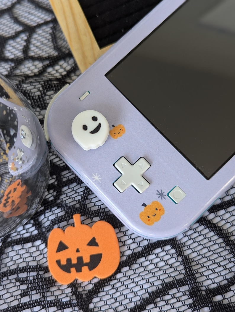 Pu&ograve; includere: Una console portatile color viola chiaro con un copri-joystick bianco a forma di fantasma, piccoli adesivi a forma di zucca arancione e un pad direzionale. Una sagoma di zucca arancione con un volto &egrave; posizionata accanto.