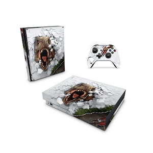 Puede incluir: Una consola Xbox One blanca y un mando con una piel que presenta un dinosaurio rompiendo una pared. La cabeza del dinosaurio es visible, con la boca abierta y los dientes a la vista.