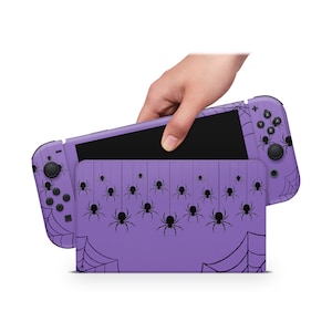 Könnte beinhalten: Lilafarbene Nintendo Switch Konsole mit einem Spinnennetz- und Spinnen-Design. Die Konsole ist geschlossen und der Bildschirm ist sichtbar.
