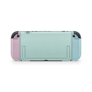 May include: A mint green and pastel colour gaming console. The console has a light blue right controller and a pink left controller. The back of the console is mint green with a black kickstand and black buttons on top.