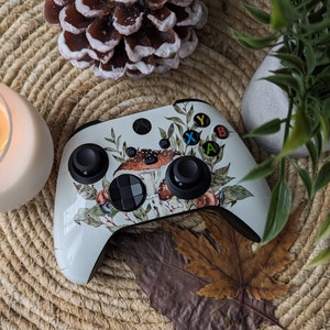 May include: A white Xbox controller with a floral and mushroom design. The controller is on a woven surface with a lit candle and a potted plant.