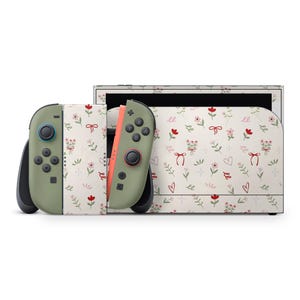 Puede incluir: Una consola y mandos Nintendo Switch con un diseño floral. Los mandos son de color verde oliva con botones negros y un lateral coral. La consola y los mandos tienen un fondo crema con flores rojas, lazos y corazones.