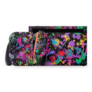 May include: A black Nintendo Switch console and Joy-Con controllers are decorated with a colorful paint splatter design. The Joy-Cons are black with pink, blue, green, and purple paint splatters. The console has a black base with the same paint splatter design.