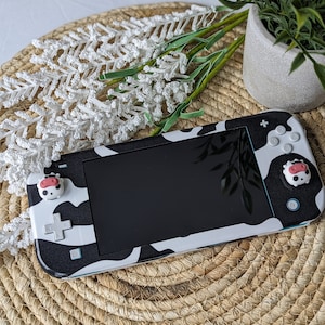以下が含まれることがあります： 黒と白の牛柄のNintendo Switch Liteで、ピンクと白の牛の形をしたボタンカバーが付いています。コンソールは、籐で編まれたマットの上に置かれています。