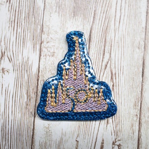 May include: A blue and pink glitter iron-on patch featuring a stylized castle with gold accents.