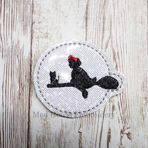 May include: A white, sparkly, round patch with a black silhouette of a witch flying on a broomstick with a black cat sitting on the broomstick. The witch is wearing a red bow.