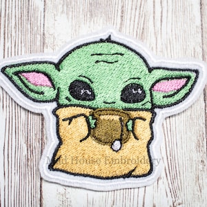 May include: Green embroidered patch of a cartoon character with large eyes, holding a cup. The character has pink ears and is wearing a brown robe.