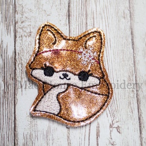 May include: A gold glitter fox with a white snowflake on its head. The fox has black eyes and a pink nose. The fox is embroidered and has a white outline.