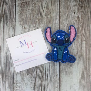 May include: Blue and pink embroidered iron-on patch of Stitch from Lilo & Stitch. The patch is sitting on a wooden surface.