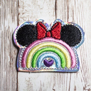 May include: A rainbow-colored embroidered patch with a red bow and black mouse ears. The patch has a glittery finish.