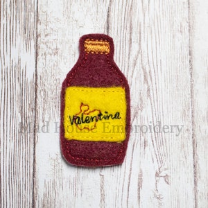 May include: A felt bottle with a yellow label that says "Valentina" in black embroidery. The bottle is maroon with a gold top.
