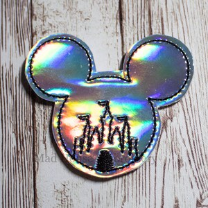 May include: A holographic Mickey Mouse head-shaped patch with a black stitched outline. The patch features a black silhouette of a castle in the center. The background is a light wood grain.