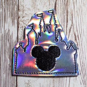 May include: A silver iridescent iron-on patch featuring a black outline of a castle with Mickey Mouse ears in the center. The text "Mad House Embroidery" is visible at the bottom of the patch.