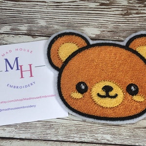 May include: Embroidered iron-on patch of a brown bear with a yellow nose and eyes. The bear has a happy expression and is surrounded by a white outline.