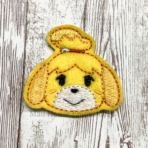 May include: A yellow felt patch featuring a cartoon dog with black eyes, a white muzzle, and a red collar. The patch has embroidered details and the text "Mad House Embroidery".