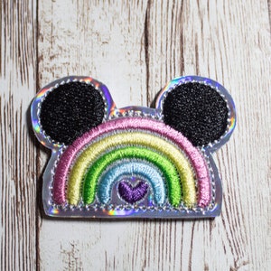 May include: A rainbow-colored embroidered patch with Mickey Mouse ears. The rainbow has a purple heart in the center. The patch is on a white wooden background.