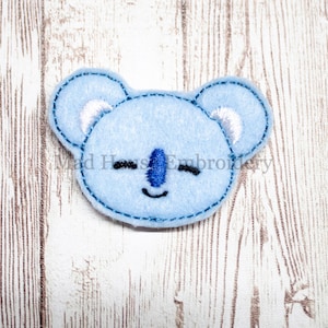 May include: A light blue koala bear applique with embroidered details. The koala has a blue nose, closed eyes, and a stitched outline. The text "Mad House Embroidery" is visible. The applique is made of felt.