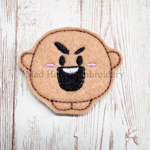 May include: A tan felt applique featuring an angry cartoon face with black eyes, eyebrows, and mouth. The applique has small ears and pink blush marks. The design is outlined with brown stitching. The text "Mad Ho...roidery" is visible.