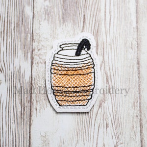 May include: Embroidered patch of a coffee drink with a black straw. The drink is brown with horizontal lines and a white top. The patch is outlined in black stitching and has a white border. The background is a light wood grain.