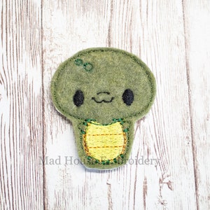 May include: A green felt turtle applique with a yellow stitched shell. The turtle has black eyes and a smiling mouth. The applique is embroidered with green thread and has the text "Mad House Embroidery".