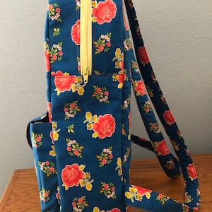 Peut inclure: Un sac à dos bleu à fleurs avec une fermeture éclair jaune et des bretelles réglables. Le sac à dos a un motif floral avec des roses rouges et de petites fleurs blanches.