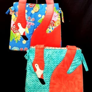 Puede incluir: Dos bolsas de tela con diseños de flamencos. La bolsa más grande tiene un fondo azul con estampado floral y un flamenco rojo. La bolsa más pequeña tiene un fondo turquesa con un patrón de chevron y un flamenco rojo.