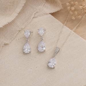 May include: A jewelry set consisting of a pair of drop earrings and a matching pendant necklace. Each piece features a clear, pear-shaped faceted stone suspended from a smaller round stone, all set in a bright silver-toned metal finish.