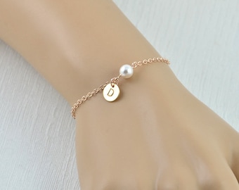 May include: A rose gold chain bracelet with a single pearl charm and a small disc charm with the letter "D" engraved on it.
