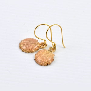 May include: A pair of gold earrings featuring a seashell design. The seashells are a light pink colour with gold accents.