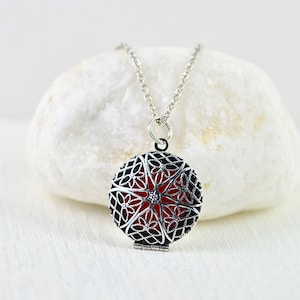 Könnte beinhalten: Silberne Halskette mit Anhänger, der ein filigranes Design und eine rote Mitte hat. Der Anhänger ist offen und das Innere ist sichtbar.