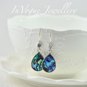 Può includere: Orecchini a goccia con intarsi in conchiglia di abalone iridescente incastonati in metallo color argento. Ogni orecchino presenta una piccola pietra rotonda trasparente sfaccettata in alto, appesa a un gancio. È visibile il testo InVogue Jewellery.