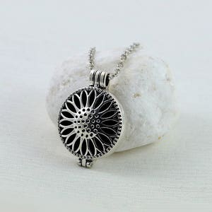 May include: A silver necklace with a round pendant featuring a detailed sunflower design. The pendant has a hinged opening, suggesting it may be a locket or aromatherapy diffuser.