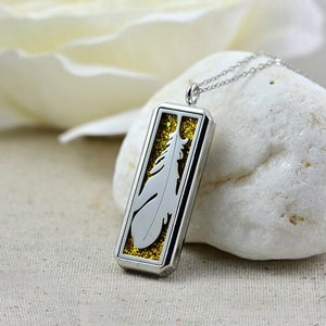 Op de afbeelding: Zilverkleurige rechthoekige hangerketting met een veermotief. De hanger heeft een zilveren frame met een uitgesneden veermotief en een gouden glitterachtergrond. De ketting wordt gepresenteerd op een witte steen.