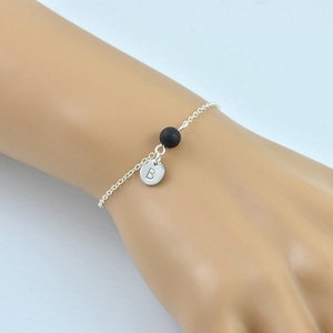 May include: A silver chain bracelet featuring a small black bead and a round, silver charm. The charm is engraved with the letter "B". The bracelet is shown on a person's wrist.