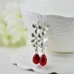 May include: Silver flower dangle earrings with red teardrop beads. The earrings are hanging from a white bowl.