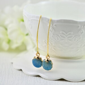 May include: A pair of gold-colored drop earrings featuring blue seashell pendants. The seashell pendants have a textured surface and are accented with gold-colored trim. The earrings are displayed on a white surface.