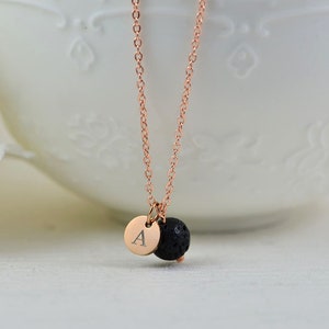 May include: A rose gold necklace featuring a small, circular pendant with the letter "A" and a black lava rock bead. The necklace is displayed against a white backdrop.