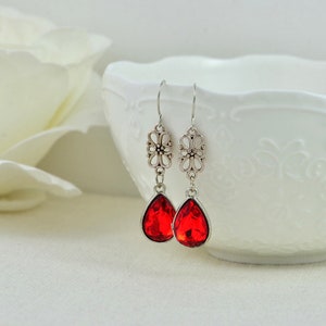 May include: A pair of silver dangle earrings with red teardrop-shaped crystals. The earrings feature a delicate floral design.