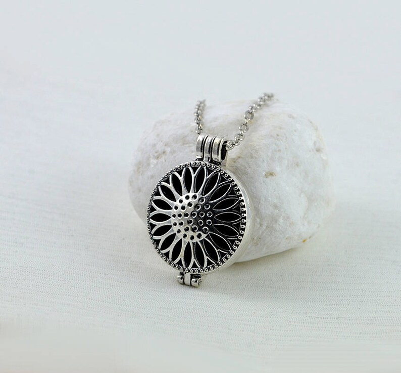 May include: A silver necklace with a round pendant featuring a detailed sunflower design. The pendant has a hinged opening, suggesting it may be a locket or aromatherapy diffuser.