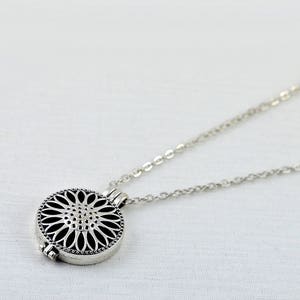 May include: A silver necklace with a round pendant featuring a delicate, intricate floral design. The pendant has a hinged opening, suggesting it may be a locket or aromatherapy diffuser.