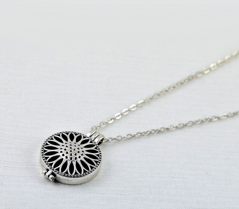 May include: A silver necklace with a round pendant featuring a delicate, intricate floral design. The pendant has a hinged opening, suggesting it may be a locket or aromatherapy diffuser.