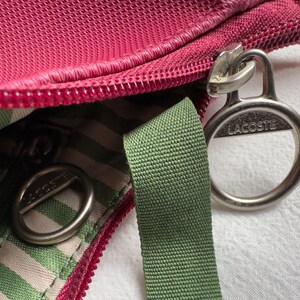 May include: Close-up of a red-pink bag with a zipper and a green strap. The bag has a striped interior and silver-colored metal hardware with the brand name "Lacoste" visible.