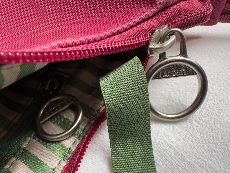 May include: Close-up of a red-pink bag with a zipper and a green strap. The bag has a striped interior and silver-colored metal hardware with the brand name "Lacoste" visible.