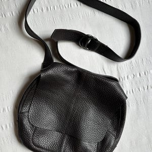 May include: Black leather crossbody bag with a textured finish. The bag features a flap closure and an adjustable strap. The strap is attached to the bag with a metal ring. The bag is sitting on a white textured surface.