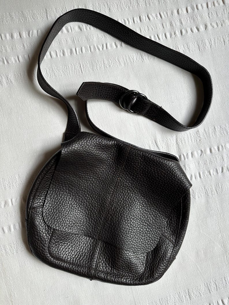 May include: Black leather crossbody bag with a textured finish. The bag features a flap closure and an adjustable strap. The strap is attached to the bag with a metal ring. The bag is sitting on a white textured surface.
