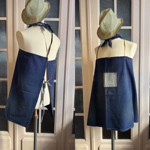 Puede incluir: Un delantal de mezclilla azul oscuro con un solo bolsillo y cierre de corbata. El delantal se muestra en un maniquí con un sombrero de paja.